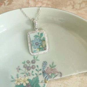 May include: A silver necklace with a rectangular pendant featuring a blue and green floral design on a white background.