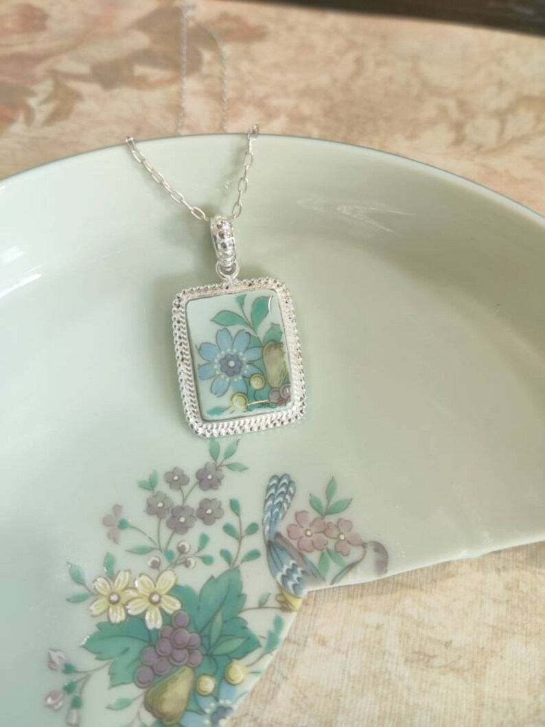 May include: A silver necklace with a rectangular pendant featuring a blue and green floral design on a white background.