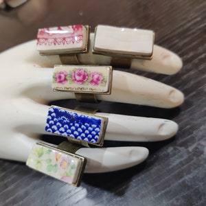 Könnte beinhalten: Fünf rechteckige Ringe mit verschiedenen Vintage-Porzellanmustern. Die Ringe sind aus Messing gefertigt und haben eine goldene Oberfläche. Die Designs umfassen Blumenmuster, ein blau-weißes Muster und einen weißen Hintergrund mit einem rot-weißen Muster.