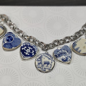 May include: A silver chain necklace with multiple blue and white porcelain heart charms. The charms feature different patterns and designs, including a buffalo with the text "Vitreous Buffalo Potter 1908".