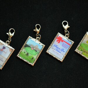 May include: Four silver charm pendants with different images. The first charm features a woman in a white dress with the text "The Earl's Intimate Enemy". The second charm has a green golf course with a building in the background. The third charm has a red and white gift box with the text "The Earl's Christmas Promise" and "Susan See Harmony". The fourth charm has a woman in a green dress with a white background.