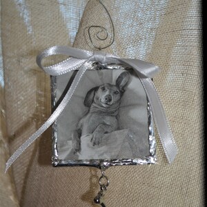 May include: A silver-framed ornament with a black and white photo of a dachshund inside. The ornament has a silver ribbon and a silver bone charm hanging from it.