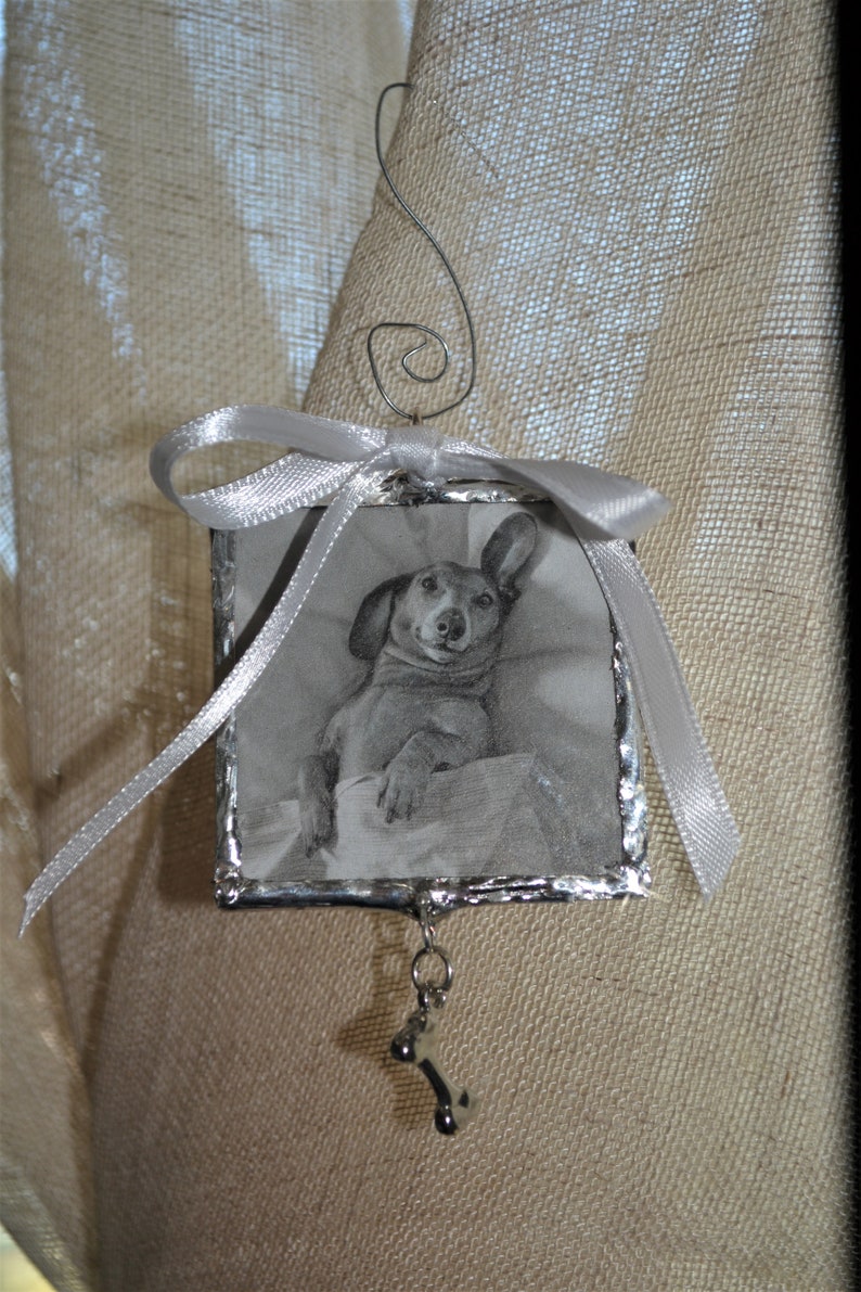 May include: A silver-framed ornament with a black and white photo of a dachshund inside. The ornament has a silver ribbon and a silver bone charm hanging from it.