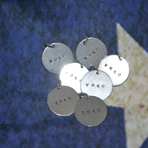 May include: A group of seven silver metal charms with the word "vote" engraved on each charm. The charms are arranged in a circle on a blue and white fabric background.