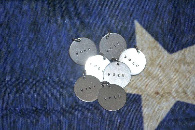 May include: A group of seven silver metal charms with the word "vote" engraved on each charm. The charms are arranged in a circle on a blue and white fabric background.