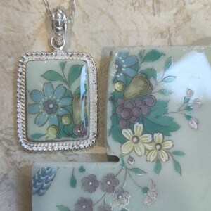 May include: A silver chain necklace with a rectangular pendant featuring a floral design on a light blue background. The pendant is surrounded by a silver border with a beaded edge. The pendant is attached to the chain with a silver clasp.