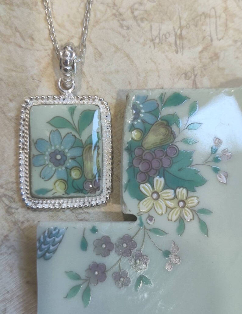 May include: A silver chain necklace with a rectangular pendant featuring a floral design on a light blue background. The pendant is surrounded by a silver border with a beaded edge. The pendant is attached to the chain with a silver clasp.
