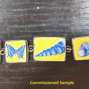 May include: A bracelet with a yellow background and blue images of seashells, a butterfly, a dragonfly, and a seashell. The bracelet is made of metal and has a clasp.