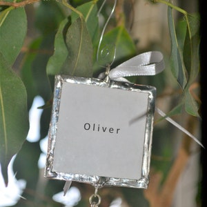 May include: Silver ornament with a white background and the name Oliver written in black. A silver bone charm hangs from the bottom.
