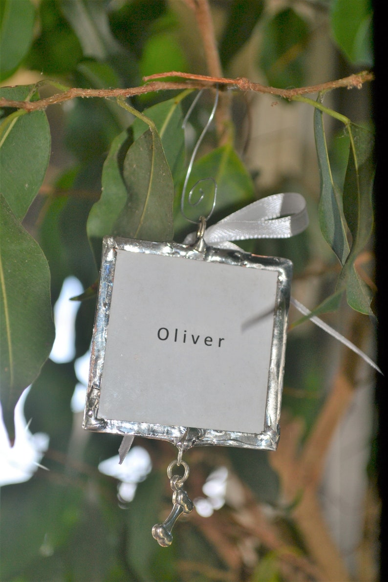 May include: Silver ornament with a white background and the name Oliver written in black. A silver bone charm hangs from the bottom.