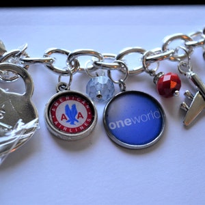 Puede incluir: Un brazalete de plata con varios dijes, incluyendo un dije de coraz&oacute;n con "925" grabado, un dije redondo con el logotipo de American Airlines, un dije redondo con el logotipo "oneworld", una cuenta roja, un dije de avi&oacute;n de plata y un dije de plata con el logotipo de American Airlines sobre un fondo azul.