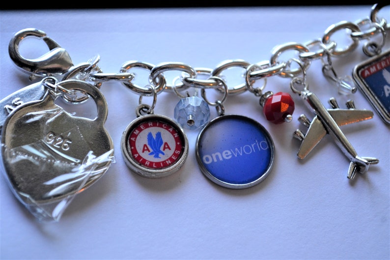 Puede incluir: Un brazalete de plata con varios dijes, incluyendo un dije de coraz&oacute;n con "925" grabado, un dije redondo con el logotipo de American Airlines, un dije redondo con el logotipo "oneworld", una cuenta roja, un dije de avi&oacute;n de plata y un dije de plata con el logotipo de American Airlines sobre un fondo azul.