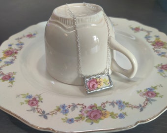 May include: A silver chain necklace with a rectangular pendant featuring a floral design in shades of pink, yellow, and white. The pendant is set in a silver frame and is resting on a white teacup.