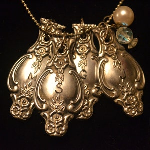 May include: A silver necklace with four antique silver spoons attached to it. The spoons have ornate designs and are connected to the chain with jump rings. The necklace also features a pearl and a crystal bead.