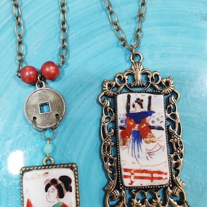 May include: Two necklaces with bronze chains and pendants. One pendant is a square with a painted image of a woman in a red and blue kimono. The other pendant is a round coin with Chinese characters. Both pendants have red beads.