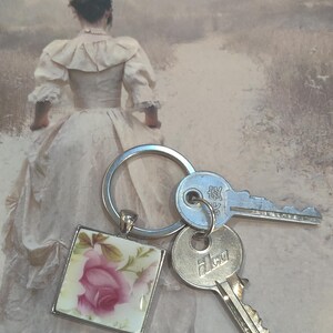 May include: A silver key chain with a square charm featuring a pink rose on a white background. The keychain also includes two silver keys, one with the word "HLCU" and the other with a small, unreadable inscription.