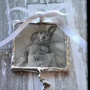 May include: A silver-framed photo ornament with a black and white image of a dog inside. The ornament has a white ribbon and a silver bone charm hanging from it.