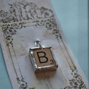 May include: A silver charm with a Scrabble tile featuring the letter "B" and the number "3" on a white background with a decorative border.