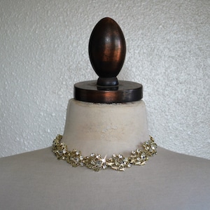May include: A gold tone choker necklace with clear rhinestones set in a floral design. The necklace is displayed on a white mannequin bust.