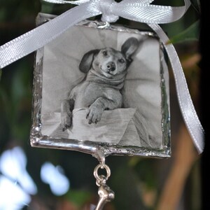 May include: A silver ornament with a black and white photo of a dachshund dog inside. The ornament has a silver bone charm hanging from it.