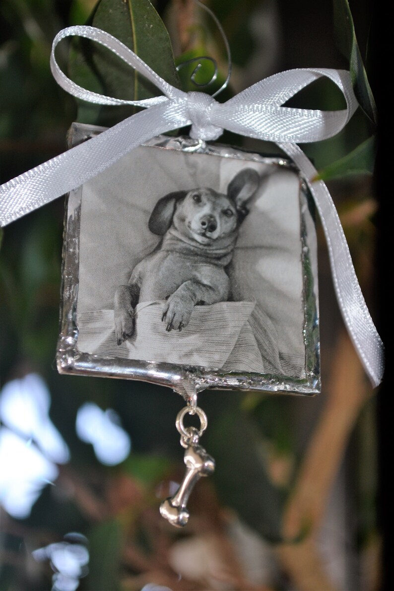 May include: A silver ornament with a black and white photo of a dachshund dog inside. The ornament has a silver bone charm hanging from it.