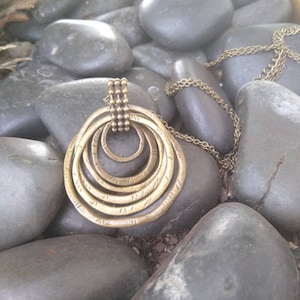 May include: A gold-toned necklace with a circular pendant made of multiple concentric rings. The pendant has a textured surface and is surrounded by smooth, grey stones.