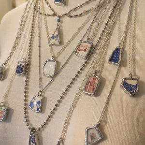 May include: A collection of necklaces with various pendants made from vintage china. The pendants are all different shapes and sizes, and they feature a variety of floral and geometric designs. The necklaces are all silver in color and are layered on a white mannequin.