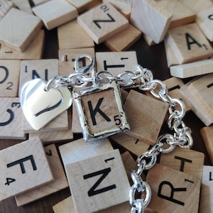 May include: A silver charm bracelet with a heart charm and a letter K charm. The charms are surrounded by wooden tiles with letters on them.