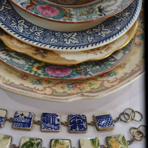 Peut inclure: Une pile de cinq assiettes en porcelaine vintage avec des motifs floraux et géométriques dans des tons de bleu, or, rose et blanc. Les assiettes sont disposées en forme de pyramide, la plus grande assiette en bas et la plus petite en haut. Un bracelet en porcelaine bleu et blanc avec des accents dorés est au premier plan.