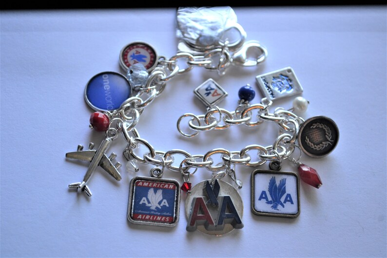 Puede incluir: Un brazalete de plata con varios abalorios, incluyendo un peque&ntilde;o avi&oacute;n, cuentas rojas y azules, y abalorios con el logotipo de American Airlines. El brazalete tambi&eacute;n tiene un abalorio con las palabras "oneworld" y un abalorio con una corona.