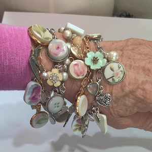 May include: A selection of charm bracelets with diverse charms. The charms include floral designs, pearls, buttons, and heart-shaped pendants. The bracelets are connected by silver-coloured chains and are adorned with a variety of colours, including pink, white, and gold.