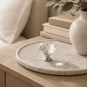 Puede incluir: Un pomo de armario de cristal transparente facetado con una base de metal plateado pulido. El pomo presenta una forma de diamante con múltiples facetas que refleja la luz, creando un aspecto brillante y centelleante en la pieza de herraje.