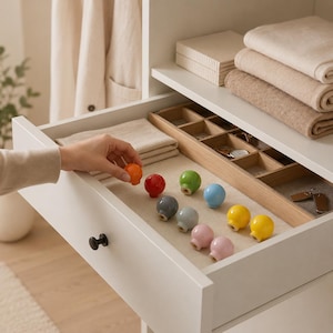 May include: A collection of small, round, solid-colored ceramic knobs in orange, red, green, blue, yellow, gray, and pink. These smooth, spherical drawer pulls are arranged inside an open white drawer next to a wooden organizer tray.