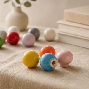 Puede incluir: Una colección de pomos esféricos para armarios en varios colores sólidos, incluyendo amarillo, azul, rosa, naranja, rojo, blanco y gris. Cada pomo presenta un acabado liso y brillante con una inserción metálica roscada en la base para su montaje.