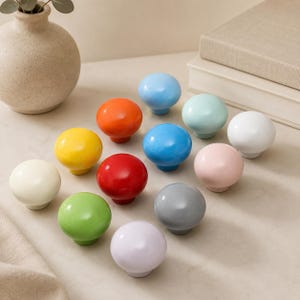 May include: A collection of twelve round, smooth cabinet knobs in a variety of solid colors including red, orange, yellow, light green, blue, light blue, white, cream, gray, pink, and lavender. Each knob features a simple, spherical design.