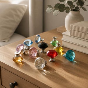 Puede incluir: Una colección de pomos para armarios con forma de diamante y facetas en varios colores, incluyendo rojo, azul, amarillo, rosa, negro y cristal transparente. Cada pomo cuenta con una base de metal plateado y una parte superior facetada que refleja la luz.