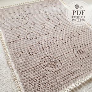 Puede incluir: Una manta rectangular de ganchillo en color beige con un patrón texturizado de un oso, corazones y el nombre AMELIE. La manta está bordeada con pequeños pompones blancos. Una marca de agua circular en la esquina dice PDF CROCHET PATTERN.