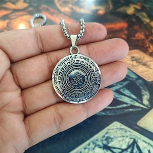 Op de afbeelding: Een zilverkleurige ronde hanger met een centraal Om-symbool omringd door ingewikkelde concentrische patronen en Sanskriet-tekst. De hanger is bevestigd aan een zilverkleurige metalen kralenketting met een gedetailleerde afwerking.