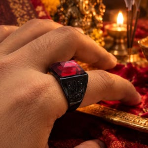 Könnte beinhalten: Ein schwarzer Metallring mit einem großen, quadratisch geschliffenen, facettierten roten Edelstein in der Mitte. Die Ringschiene weist an den Seiten komplizierte, dunkel gravierte Muster auf, die einen strukturierten Kontrast zur glatten Oberfläche bilden.
