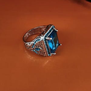 Könnte beinhalten: Ein silberfarbener Ring mit einem großen, rechteckigen, facettierten blauen Mittelstein, der von vier Krappen gehalten wird. Die Ringschiene ist mit filigranen Metallverzierungen und kleinen, runden, hellblauen Akzentsteinen besetzt.
