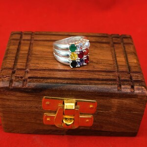 Può includere: Un anello in argento con un gruppo quadrato di nove piccole pietre preziose multicolori, tra cui pietre verdi, gialle, nere, bianche, rosse e blu, montato su un'ampia fascia in argento lucido con una finitura liscia e riflettente.