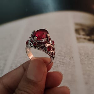 Könnte beinhalten: Ein silberfarbener Ring mit einem großen, oval geschliffenen, tiefroten Mittelstein. Das Metallband ist mit filigranen Mustern verziert und mit kleineren, runden roten Steinen besetzt, die entlang der Seiten der Fassung eingelassen sind.