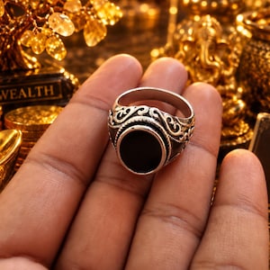 Könnte beinhalten: Ein silberfarbener Ring mit einem glatten, kreisförmigen schwarzen Steineinsatz in der Mitte. Das Band ist mit komplizierten, erhabenen Schnörkelmustern verziert, die von den Seiten der zentralen Fassung ausgehen und ein strukturiertes Aussehen verleihen.