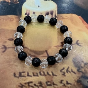 Op de afbeelding: Een cirkelvormige kralenarmband met een afwisselend patroon van gladde, ondoorzichtige zwarte bolvormige kralen en heldere, gefacetteerde, transparante kristalachtige kralen. De kralen zijn in een doorlopende lus geregen met een hoog contrast.