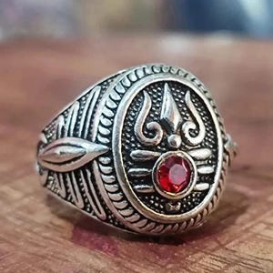 May include: A silver-toned metal ring featuring an oval face with an embossed trident symbol and a small, round, faceted red stone at the base. The band and the sides of the ring display intricate, textured, and raised linear decorative patterns.