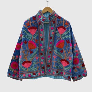 May include: A blue jacket featuring vibrant floral embroidery in shades of pink, orange, and green. The garment has a stand-up collar, long sleeves, and an open front design. The interior lining is visible and displays a light blue and white striped pattern.