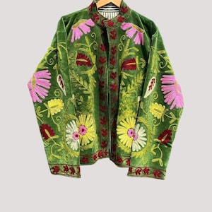 Peut inclure: Une veste verte à manches longues ornée de broderies florales aux tons rose, jaune et rouge foncé. Le vêtement présente un col montant, une ouverture frontale et une doublure rayée noire et blanche visible au niveau de l'encolure. Broderies sur le devant.