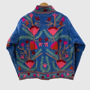 May include: A blue jacket featuring vibrant floral embroidery in shades of pink, red, and green. The design includes symmetrical botanical patterns with large stylized flowers and leafy stems covering the back and sleeves, finished with a high collar.