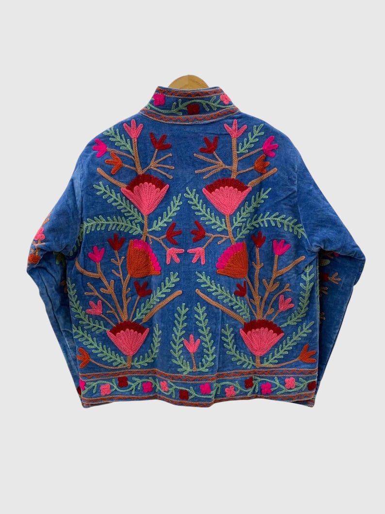 May include: A blue jacket featuring vibrant floral embroidery in shades of pink, red, and green. The design includes symmetrical botanical patterns with large stylized flowers and leafy stems covering the back and sleeves, finished with a high collar.