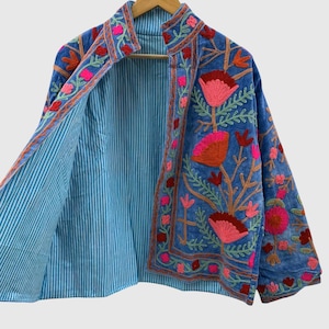 May include: A blue jacket featuring vibrant floral embroidery in shades of pink, red, and orange. The garment has a stand-up collar and long sleeves, with an interior lining that displays a light blue and white vertical pinstripe pattern.